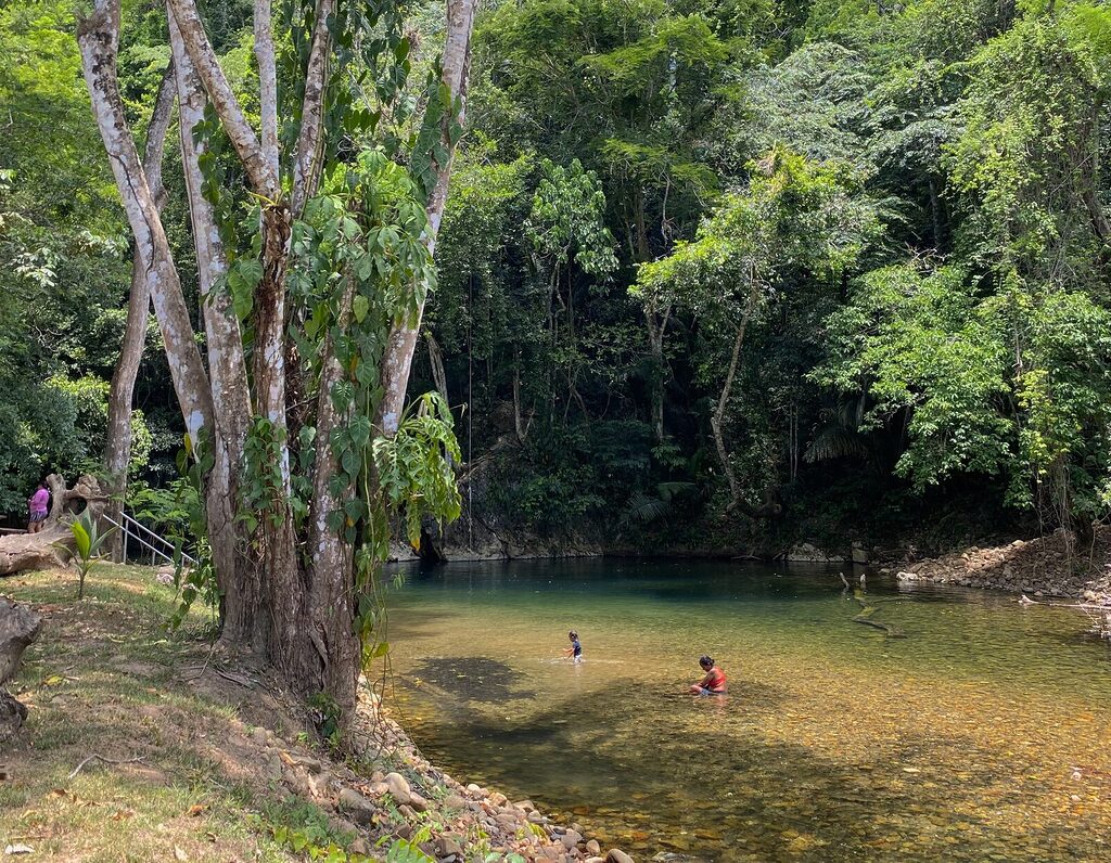 A serene natural swimming area at Barton Creek Unique Resort in Belize, featuring crystal-clear water, lush jungle surroundings, and a shaded picnic table—ideal for families and nature lovers.