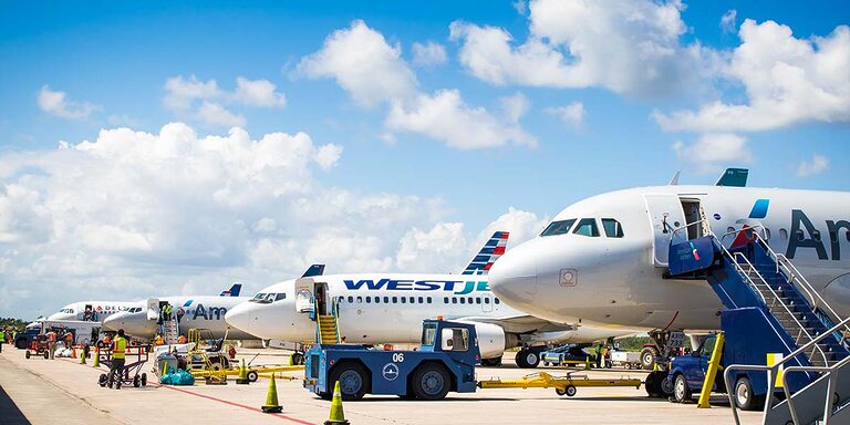 Travelers disembarking on the tarmac under blue skies at Belize International Airport, with open airplane doors and ground crew assisting. (Flights to Belize)