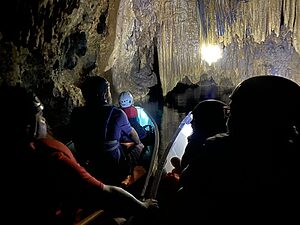 A canoe pauses under a low limestone formation inside Barton Creek Cave in Belize, with light illuminating the rock and people seated quietly inside.