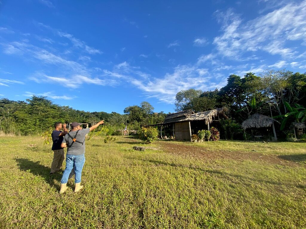 A man uses binoculars while a Belizean guide points toward birds in the trees behind thatched-roof buildings, under a blue sky with green grass in August.