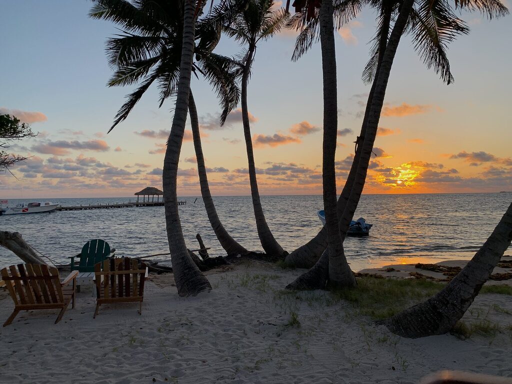 Sunrise over Long Caye near Belize’s Blue Hole in September, viewed between two coconut trees with a dock and beach chairs in the distance.