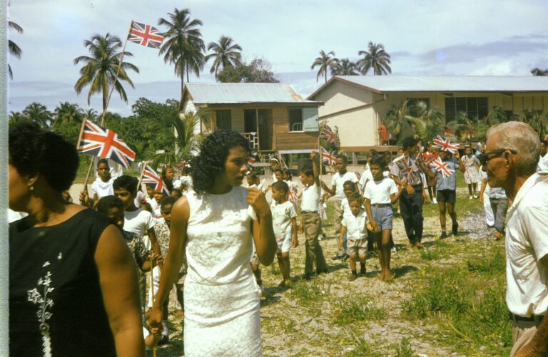 Children and adults in Placencia, Belize during Queen of the Bay Pageant, 1965–1967, with British flag during St. George’s Caye Day