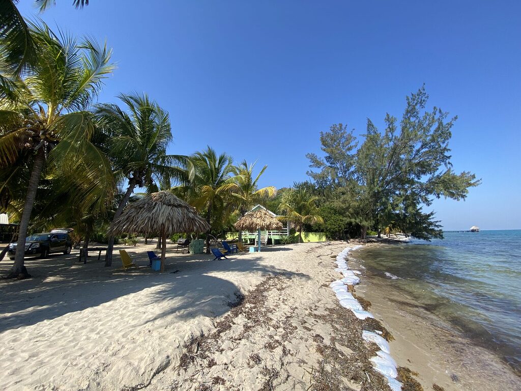 Sandbags line the beachfront at Village Inn in Placencia, Belize, with a cabana, coconut trees, and clear blue sky in April.