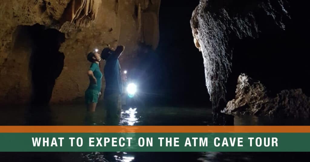 Visitors exploring the ATM Cave in Belize with headlamps, standing in shallow water beneath stalactites.