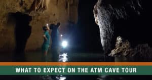 Visitors exploring the ATM Cave in Belize with headlamps, standing in shallow water beneath stalactites.